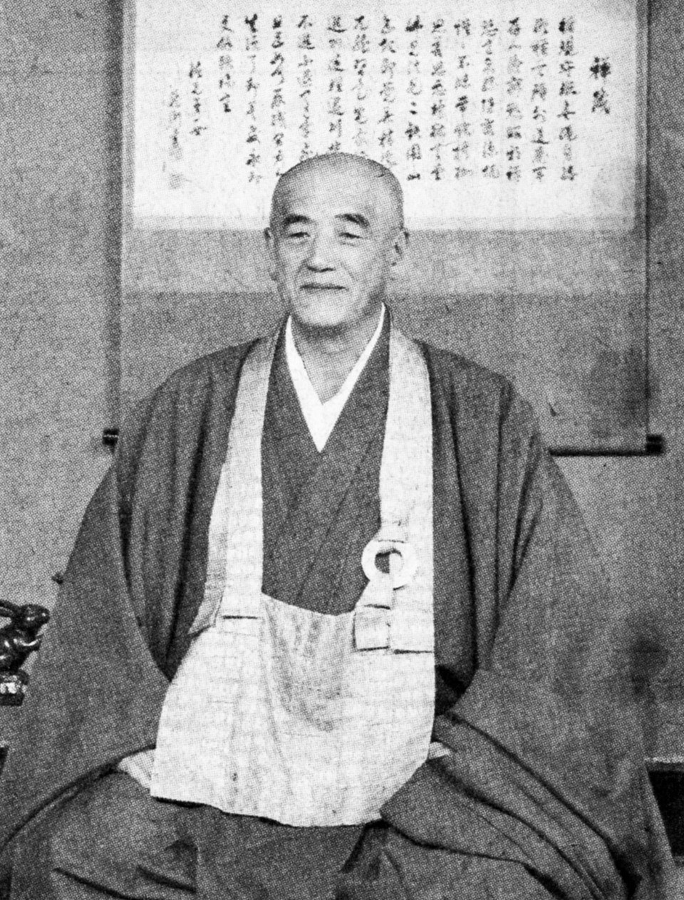 een man in een Japans monniksgewaad zit in meditatiehouding voor een scroll met calligrafie, hij kijkt vriendelijk naar de camera